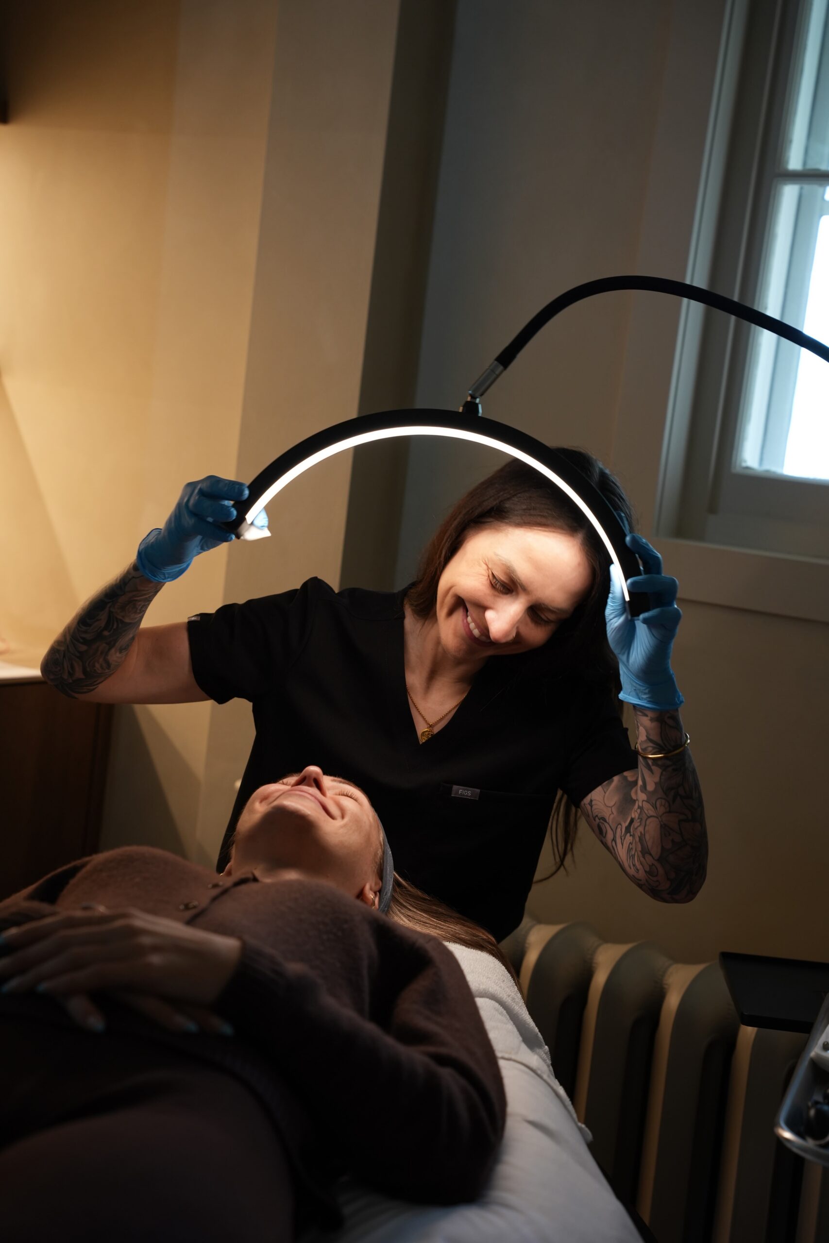An aestethician stands above a patient laying down in a bed as she adjusts the light in the medspa room