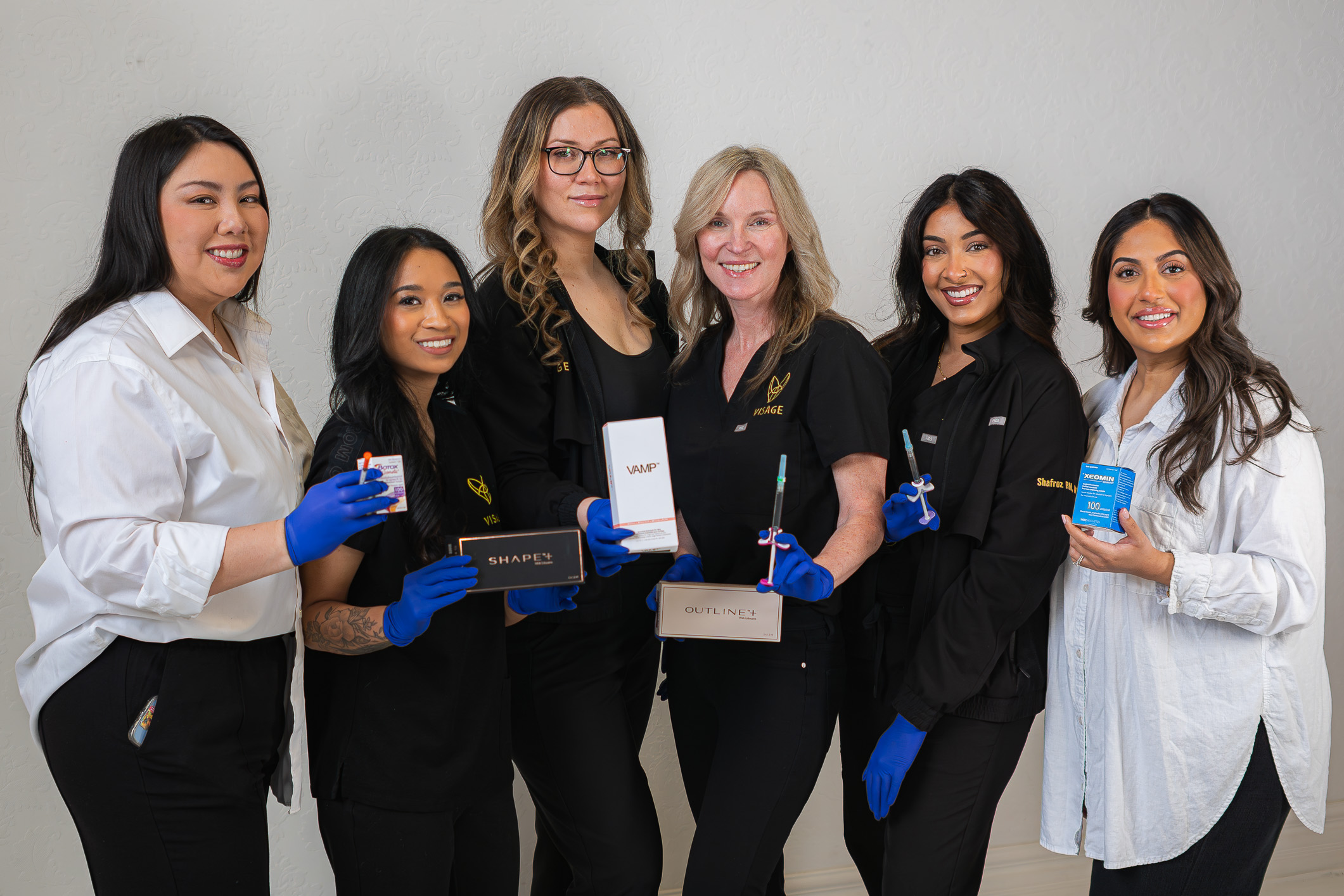 Group of 6 female nurse injectors stand side by side in front of a white backdrop each holding a medical aesthetic products